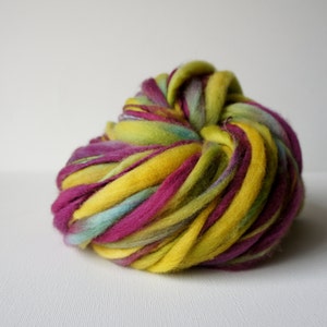 May include: A vibrant ball of hand-dyed merino wool roving. The wool features bold colors including yellow, magenta, teal, and green. The thick yarn is tightly wound, showcasing the rich color variations. Ideal for fiber art projects.