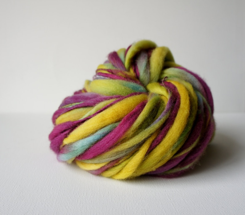 May include: A vibrant ball of hand-dyed merino wool roving. The wool features bold colors including yellow, magenta, teal, and green. The thick yarn is tightly wound, showcasing the rich color variations. Ideal for fiber art projects.