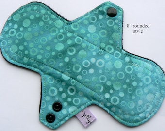 May include: An 8-inch rounded style reusable cloth menstrual pad. The pad features a teal fabric with a pattern of overlapping circles in shades of teal and green. It has black snaps and a black backing. The text "zillye" is on a small purple tag.