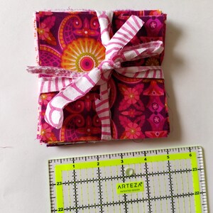 May include: A stack of patterned fabric squares in shades of pink, purple, and orange, tied with a white and pink striped ribbon. A yellow and black ruler is in the foreground.