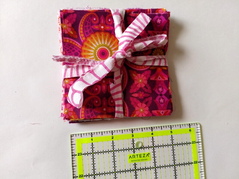 May include: A stack of patterned fabric squares in shades of pink, purple, and orange, tied with a white and pink striped ribbon. A yellow and black ruler is in the foreground.