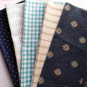 May include: A collection of folded fabric swatches in various patterns. Includes navy blue with white stars, white with gray dots and lines, teal gingham, cream with thin stripes, and navy with floral designs.