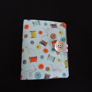 May include: A light blue fabric pouch with a button closure. The pouch is decorated with a pattern of colourful spools of thread and buttons.