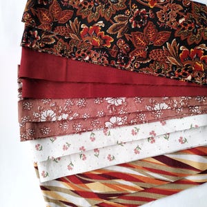 May include: A selection of fabric swatches in a range of patterns and colours. The top swatch displays a dark floral design with red and orange accents. Other swatches include solid red, floral, and striped patterns. The fabrics are layered on a white surface.