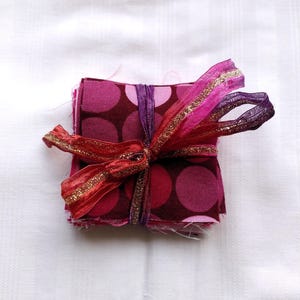 May include: A stack of fabric squares with a polka dot pattern in shades of pink and brown, tied together with a red, pink, and gold ribbon.