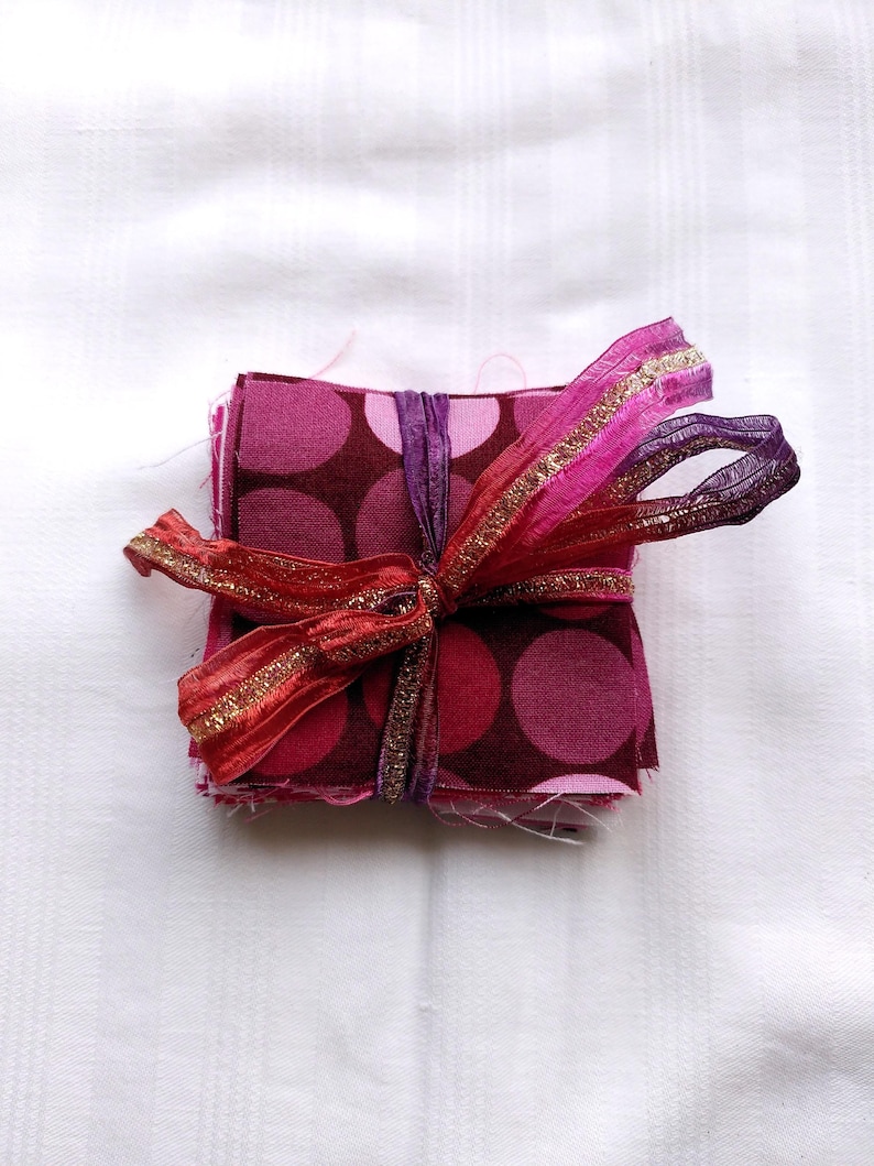 May include: A stack of fabric squares with a polka dot pattern in shades of pink and brown, tied together with a red, pink, and gold ribbon.