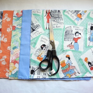 May include: A collection of six fabric scraps with various patterns, including a blue and white floral design, a red and white geometric design, a green and white travel-themed design, and a blue and white striped design. A pair of silver scissors is placed on top of the fabric scraps.