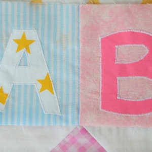 Puede incluir: Primer plano de dos bloques de tela. La letra "A" es blanca con estrellas amarillas sobre un fondo a rayas azules y blancas. La letra "B" es rosa sobre un fondo rosa. Las letras son parte de un edredón más grande.