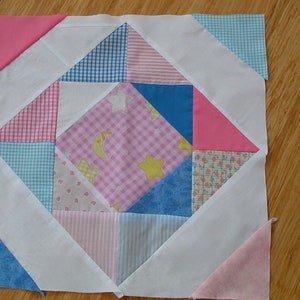 Può includere: Un blocco di trapunta patchwork con uno sfondo bianco e una varietà di tessuti a motivi rosa, blu e bianchi. Il blocco presenta un quadrato centrale con un motivo a quadri rosa e bianco e stelle, circondato da una forma a diamante composta da quadrati e triangoli più piccoli.