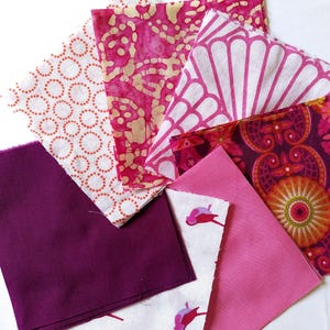 May include: A collection of colorful fabric squares in various shades of pink, purple, and white. The fabrics feature diverse patterns, including floral, geometric, and abstract designs, suitable for quilting or crafting projects.