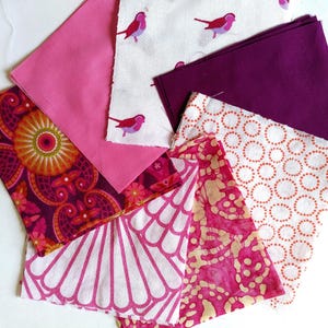 May include: A collection of fabric swatches in various patterns and colors. Includes pink, purple, and white fabrics with floral, bird, and geometric designs. The fabrics are arranged on a white surface.