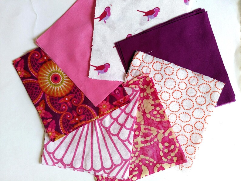 May include: A collection of fabric swatches in various patterns and colors. Includes pink, purple, and white fabrics with floral, bird, and geometric designs. The fabrics are arranged on a white surface.