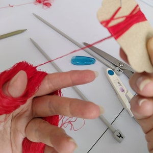 May include: A close-up of a hand holding red yarn, with two knitting needles, a pair of scissors, and a pregnancy test on a white surface.