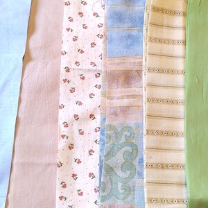 May include: A selection of fabric swatches in different colours and patterns. Includes solid light blue, pale pink, floral print with red flowers, blue and brown patterned, striped beige, and solid green fabrics. Suitable for quilting or sewing.