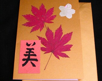 Puede incluir: Una tarjeta dorada con un cuadrado de papel rojo y negro con el carácter japonés "美" (pronunciado "bi") que significa "belleza". La tarjeta está decorada con hojas de arce rojas prensadas y una flor blanca.