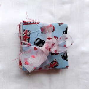 May include: A stack of fabric squares with a blue and red Union Jack pattern and black cars. The fabric squares are tied together with a pink and white ribbon.
