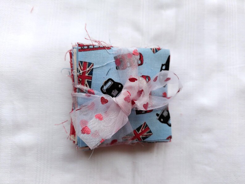 May include: A stack of fabric squares with a blue and red Union Jack pattern and black cars. The fabric squares are tied together with a pink and white ribbon.