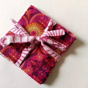 May include: A square stack of fabric squares tied with a white and pink striped ribbon. The fabric has a floral and paisley pattern in shades of pink, orange, and purple. The ribbon is tied in a bow.