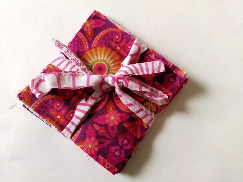 May include: A square stack of fabric squares tied with a white and pink striped ribbon. The fabric has a floral and paisley pattern in shades of pink, orange, and purple. The ribbon is tied in a bow.