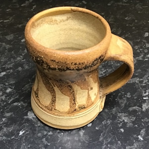 Puede incluir: Taza de cerámica hecha a mano con cuerpo redondeado y asa resistente. La taza presenta una base beige con patrones verticales marrones y beige y un borde marrón oscuro. El interior es de color beige claro.