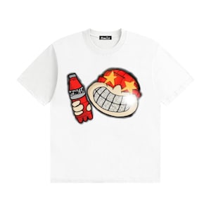 May include: A white short-sleeve t-shirt featuring a graphic of a cartoon character with a red hat, yellow star eyes, and a diamond-textured grill smile, holding a red bottle labelled Smile. The inner neck label also displays the text Smile.