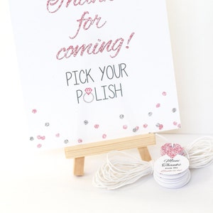 May include: A white sign with pink glitter accents reads "Thanks for coming! Pick your polish" with a pink diamond ring icon. The sign is on a small wooden easel. There are small round white tags with pink glitter accents that say "Mani Thanks from the Bride-to-be".