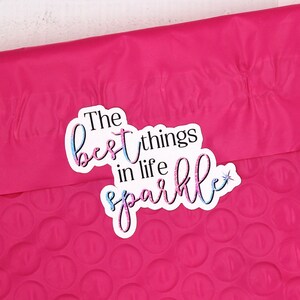 Op de afbeelding: Een vel stickers met de tekst "The best things in life sparkle" gedrukt in roze en blauwe letters met een ster. De stickers zijn op een witte achtergrond.