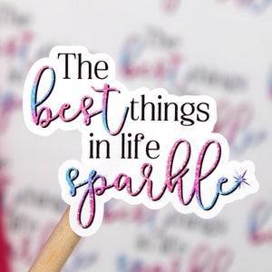 Op de afbeelding: Een sticker met roze en blauwe glitterletters met de tekst "The best things in life sparkle" met een kleine ster rechts van het woord sparkle.