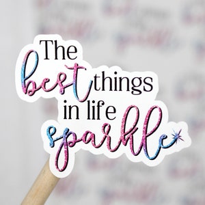 Op de afbeelding: Een roze en blauwe glitter sticker met de tekst "The best things in life sparkle" en een kleine ster.