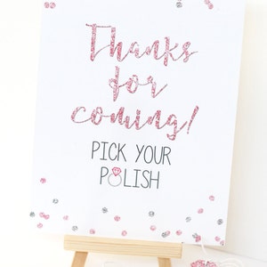 May include: A white sign with pink glitter text that says "Thanks for coming! Pick your polish" with a pink diamond ring icon. The sign is on a small wooden easel. There are pink and silver glitter confetti on the sign. A stack of small round white tags with pink glitter text that says "mant thanks from the bride-to-be!" is on a white string.