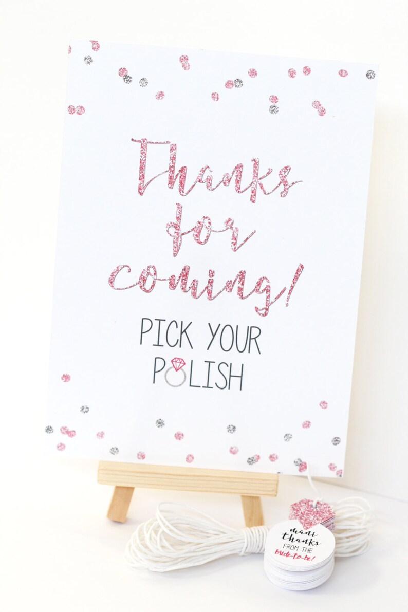 May include: A white sign with pink glitter text that says "Thanks for coming! Pick your polish" with a pink diamond ring icon. The sign is on a small wooden easel. There are pink and silver glitter confetti on the sign. A stack of small round white tags with pink glitter text that says "mant thanks from the bride-to-be!" is on a white string.