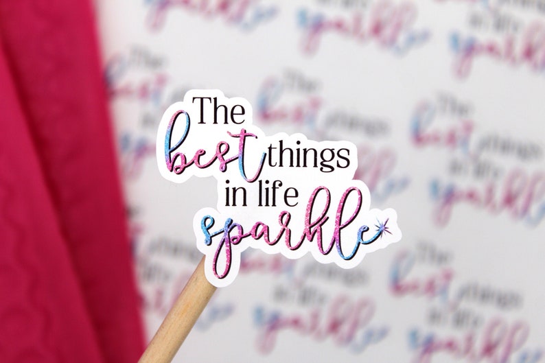 Op de afbeelding: Een sticker met roze en blauwe glitterletters met de tekst "The best things in life sparkle" met een kleine ster rechts van het woord sparkle.