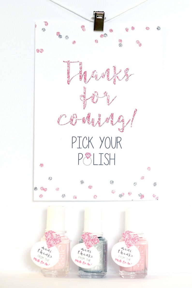 May include: A white card with pink glitter text that says "Thanks for coming! Pick your polish" with a pink diamond ring icon. Three small bottles of nail polish with pink glitter heart shaped tags that say "Made thanks from the bride-to-be".