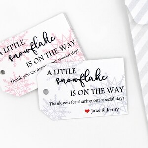 以下が含まれることがあります： 雪の結晶のデザインと「A little snowflake is on the way.」というテキストが書かれた白いギフトタグが2つあります。1つのタグにはピンクの雪の結晶が、もう1つのタグには灰色の雪の結晶が描かれています。灰色の雪の結晶が描かれたタグには、「Thank you for sharing our special day! Jake & Jenny」というテキストも書かれています。