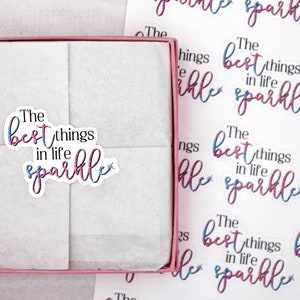 Op de afbeelding: Een roze doos met een witte tissuepapier binnenkant. Het tissuepapier heeft een sticker met de tekst "The best things in life sparkle" in roze en blauwe letters met een kleine ster.