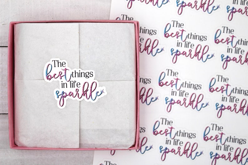 Op de afbeelding: Een roze doos met een witte tissuepapier binnenkant. Het tissuepapier heeft een sticker met de tekst "The best things in life sparkle" in roze en blauwe letters met een kleine ster.