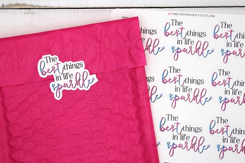 Op de afbeelding: Een vel stickers met de tekst "The best things in life sparkle" gedrukt in roze en blauwe letters met een ster. De stickers zijn op een witte achtergrond.