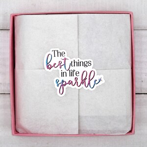 Op de afbeelding: Een roze cadeaudoos met een witte tissuepapier voering. De doos heeft een sticker met de tekst "The best things in life sparkle" met een glittereffect.
