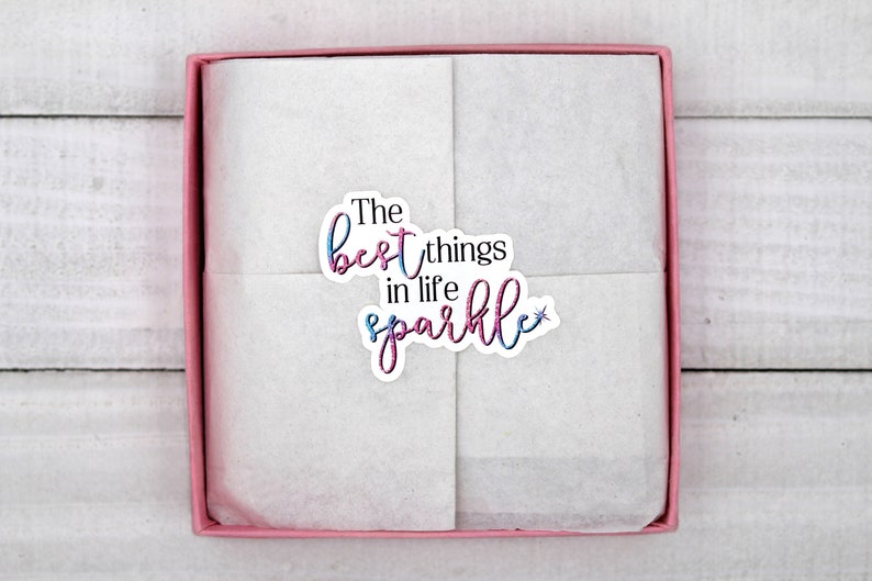 Op de afbeelding: Een roze cadeaudoos met een witte tissuepapier voering. De doos heeft een sticker met de tekst "The best things in life sparkle" met een glittereffect.