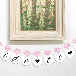 May include: Pink and white bridal shower banner with the words "Bride to Be" in black lettering. The banner is strung with pink glitter diamond shapes.