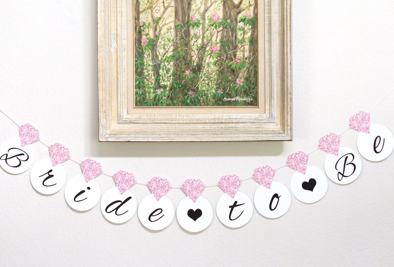 May include: Pink and white bridal shower banner with the words "Bride to Be" in black lettering. The banner is strung with pink glitter diamond shapes.