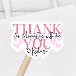 May include: Pink and white thank you sticker with the text "THANK YOU for celebrating with me!" and the name "Kaleigh" in cursive. The sticker has a heart design and is on a wooden stick.