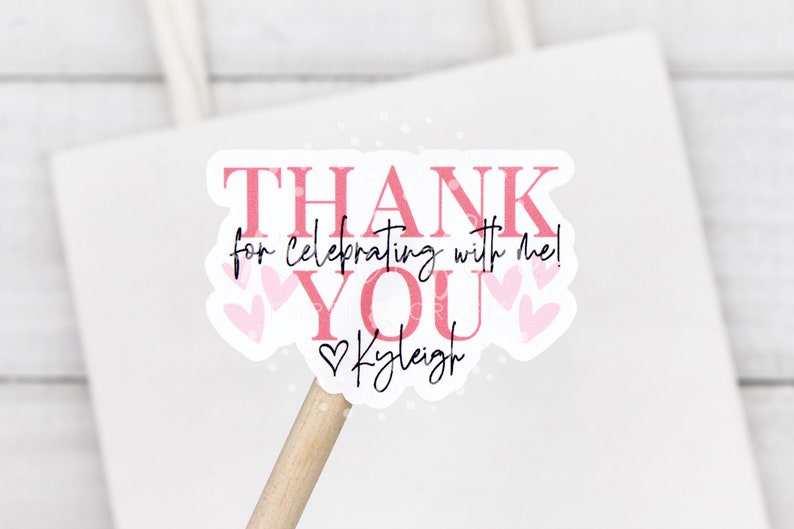 May include: Pink and white thank you sticker with the text "THANK YOU for celebrating with me!" and the name "Kaleigh" in cursive. The sticker has a heart design and is on a wooden stick.