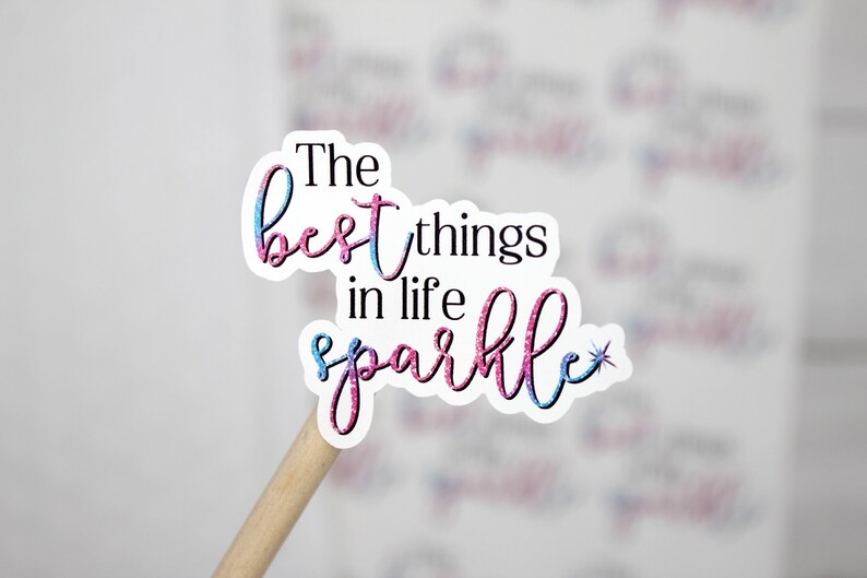 Op de afbeelding: Een roze en blauwe glitter sticker met de tekst "The best things in life sparkle" en een kleine ster.