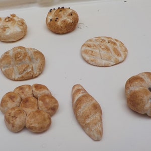 Może przedstawiać: Zestaw ośmiu różnych rodzajów bułek, w tym okrągła bułka z wzorem w kratkę, okrągła bułka z wzorem spiralnym i długa bułka ze spiczastym końcem.