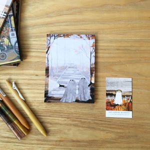 Può includere: Su una superficie di legno sono esposti articoli di cancelleria, tra cui penne e due biglietti illustrati. I biglietti presentano illustrazioni di fantasmi, uno con una scena di foresta e l'altro con zucche. Il testo "KATHERINE BLOWER ILLUSTRATOR & DESIGNER" è visibile.