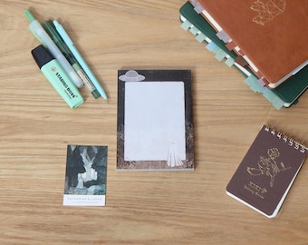 May include: A collection of stationery items on a wooden surface. Includes a notepad with a ghost and UFO design, a small notebook with a drawing, pens, a highlighter, and several journals with tabs.