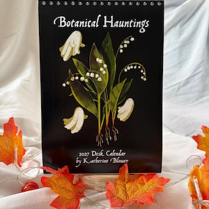 Puede incluir: Un calendario de escritorio de espiral negro titulado Botanical Hauntings 2027 Desk Calendar by Katherine Blower. La portada presenta una ilustración de lirios de los valles con hojas verdes y tres pequeñas figuras de fantasmas blancos.