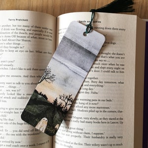 May include: A rectangular paper bookmark featuring a watercolor-style landscape with a dark, cloudy sky, a grassy hill, bare trees, and two small white ghost figures at the bottom, finished with a dark green string and black tassel.