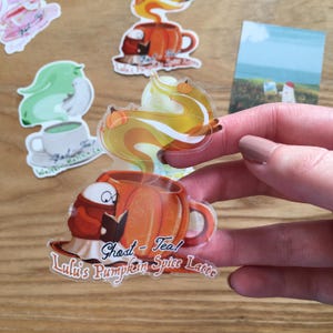 Puede incluir: Una colección de pegatinas caprichosas con fantasmas de dibujos animados y tazas de café. Una pegatina muestra un fantasma leyendo un libro dentro de una taza de café con leche de calabaza y especias. El texto de la pegatina dice "Ghost-Tea! Lulu's Pumpkin Spice Latte."